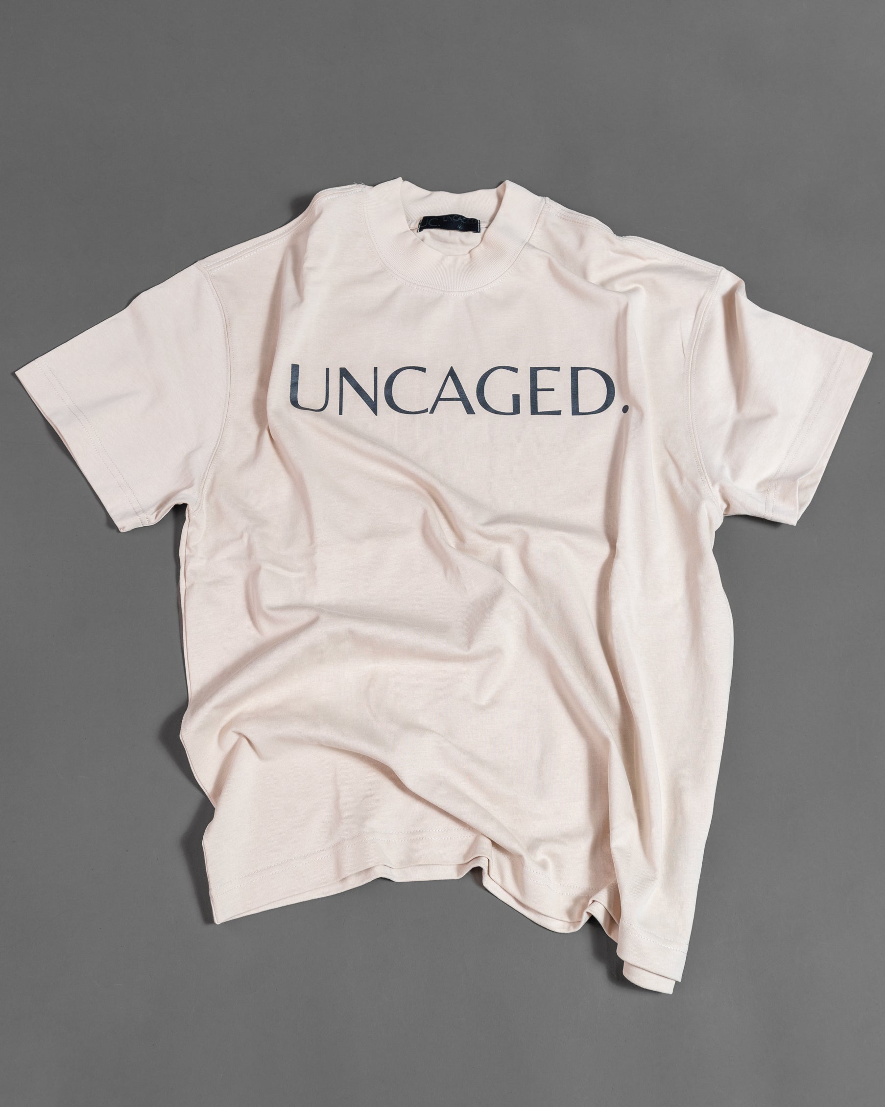 UNCAGED. Origin Tee in beige cream — heavyweight oversized fit, flat lay, tonal logo print.