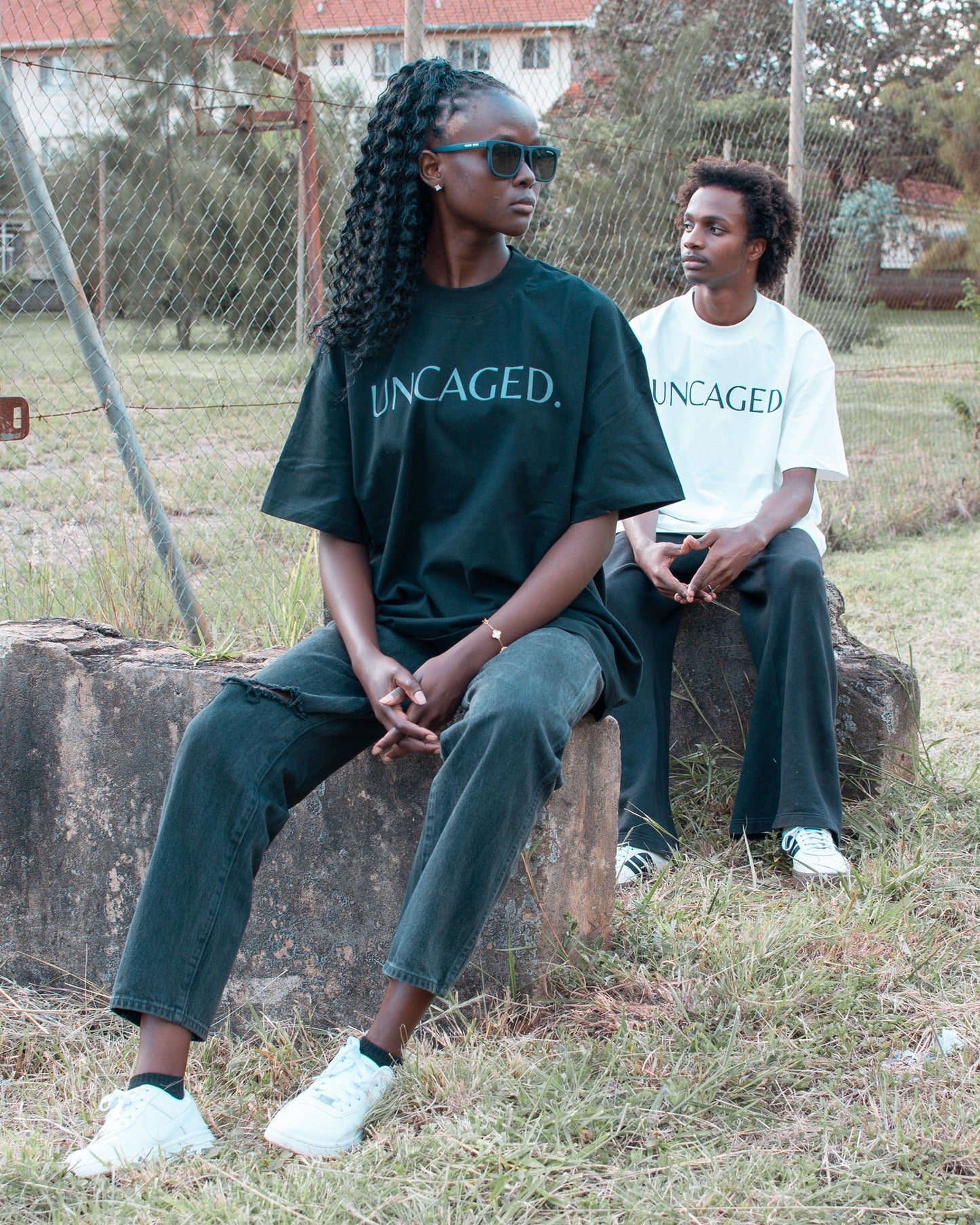 Two models wearing the UNCAGED. Origin Tee — midnight black and off-white colorways, heavyweight oversized fit with tonal logo print.