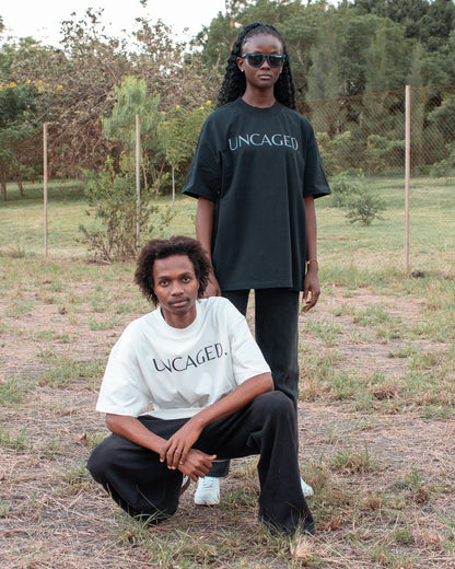 Two models wearing the UNCAGED. Origin Tee — off-white and black colorways, heavyweight oversized fit with tonal logo print.