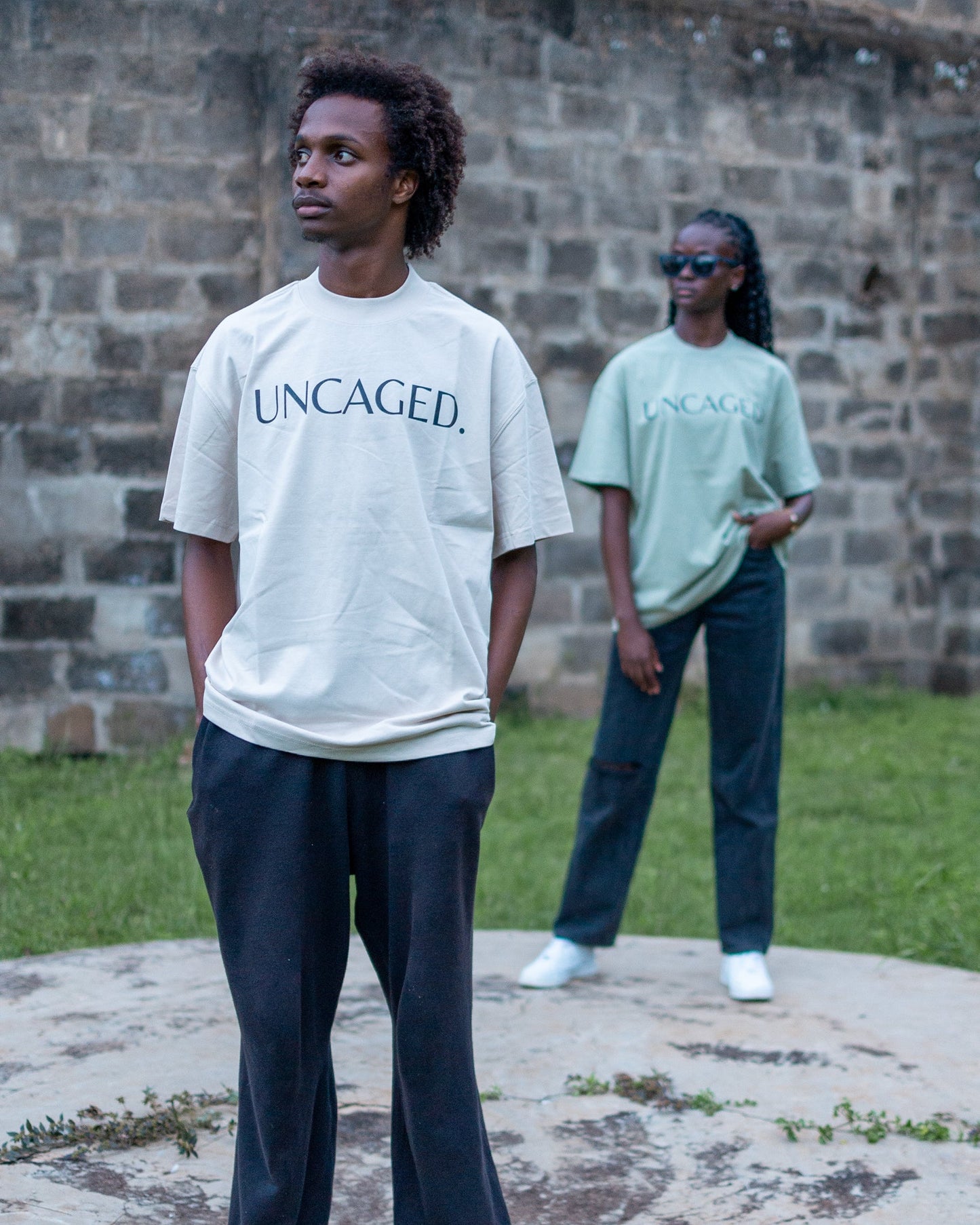 Two models wearing the UNCAGED. Origin Tee — beige cream and sage green colorways, heavyweight oversized fit with tonal logo print.