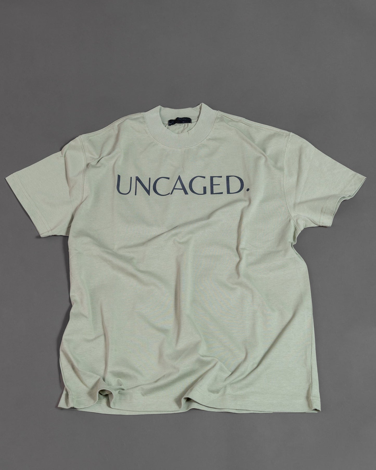 UNCAGED. Origin Tee in sage green, heavyweight oversized fit, flat lay, tonal logo print.