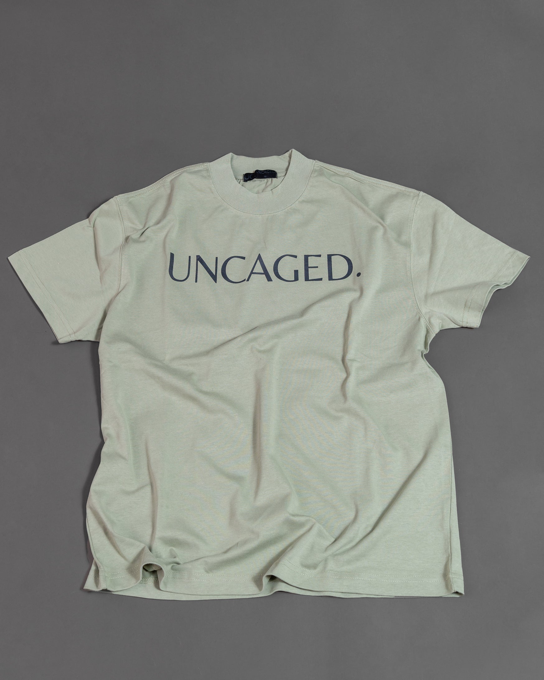 UNCAGED. Origin Tee in sage green, heavyweight oversized fit, flat lay, tonal logo print.