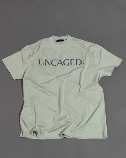 UNCAGED. Origin Tee in sage green, heavyweight oversized fit, flat lay, tonal logo print.