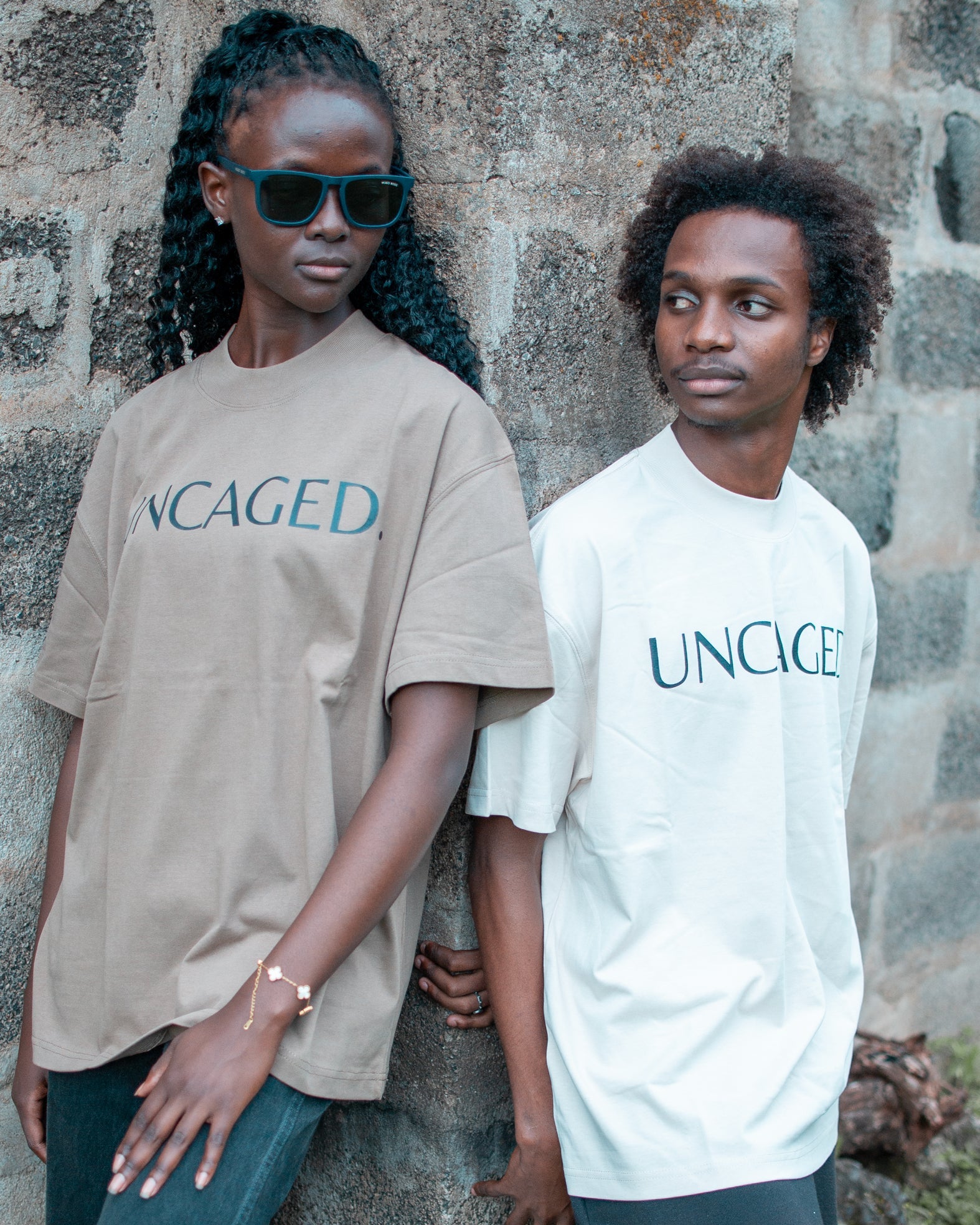 Two models wearing the UNCAGED. Origin Tee — taupe brown and beige cream colorways, heavyweight oversized fit with teal logo print