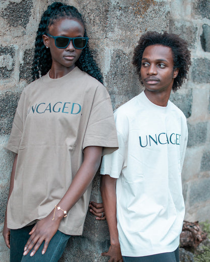 Two models wearing the UNCAGED. Origin Tee — taupe brown and beige cream colorways, heavyweight oversized fit with teal logo print