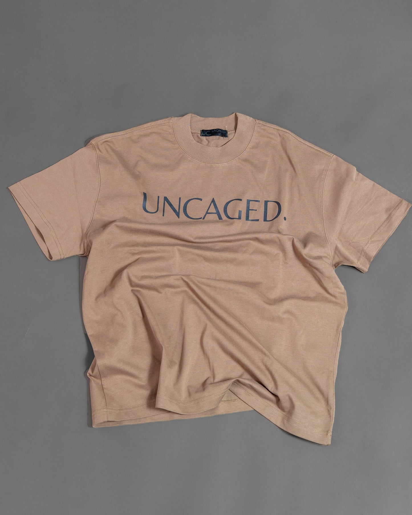 UNCAGED. Origin Tee in taupe brown — heavyweight oversized fit, flat lay, teal logo print.