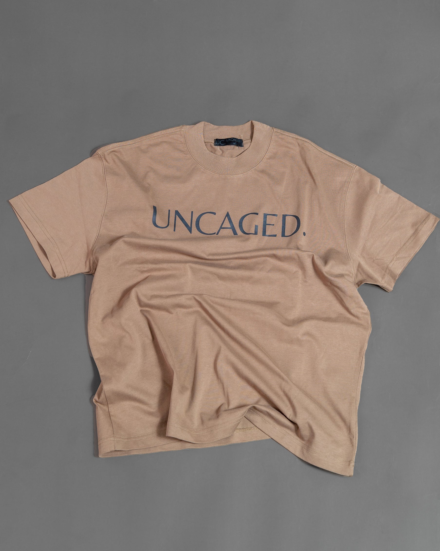 UNCAGED. Origin Tee in taupe brown — heavyweight oversized fit, flat lay, teal logo print.